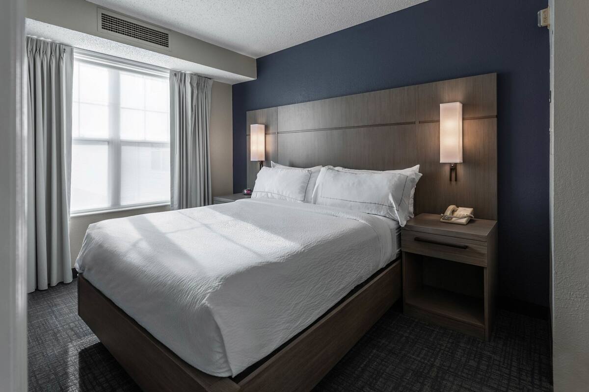 A hotel room with a large bed, two bedside tables, a headboard with integrated lighting, and a window with curtains.