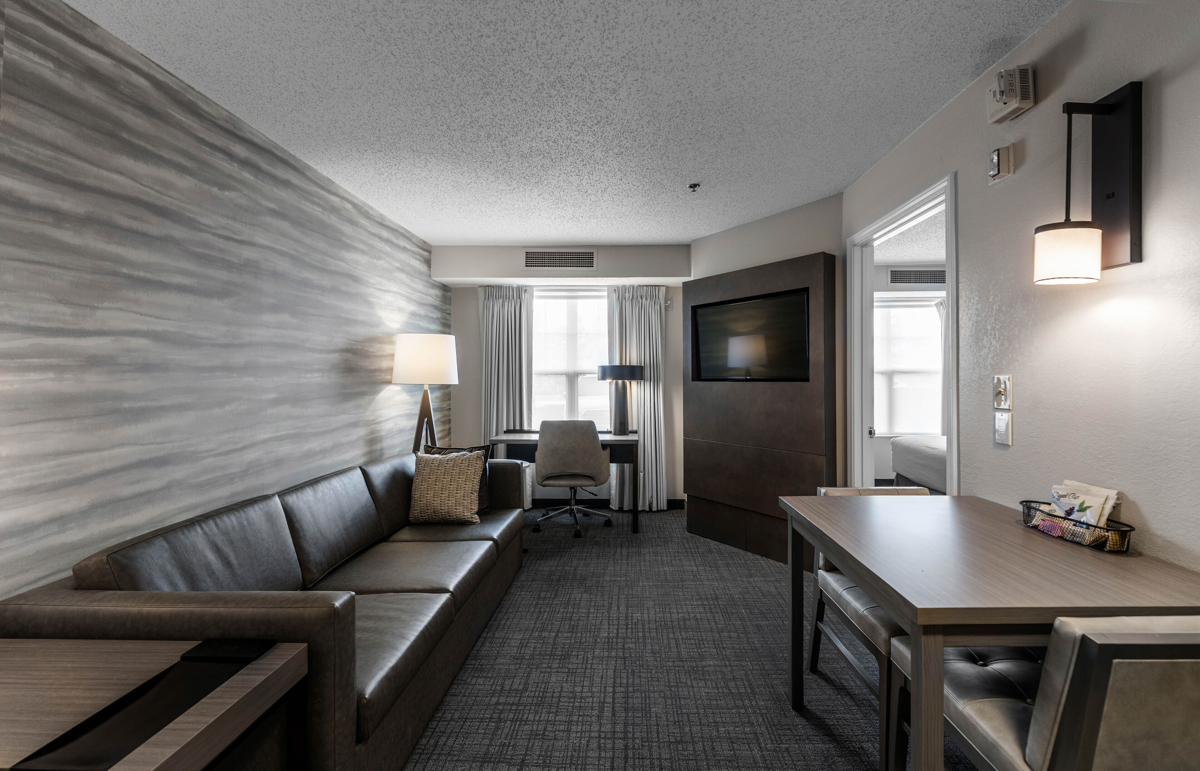 A modern hotel room with a sofa, a desk, a chair, a TV, and a lamp.
