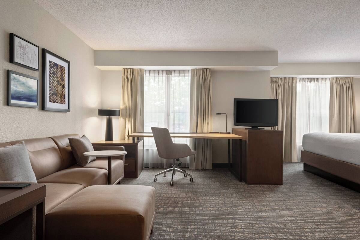 A modern hotel room with a sofa, desk, chair, TV, and bed.