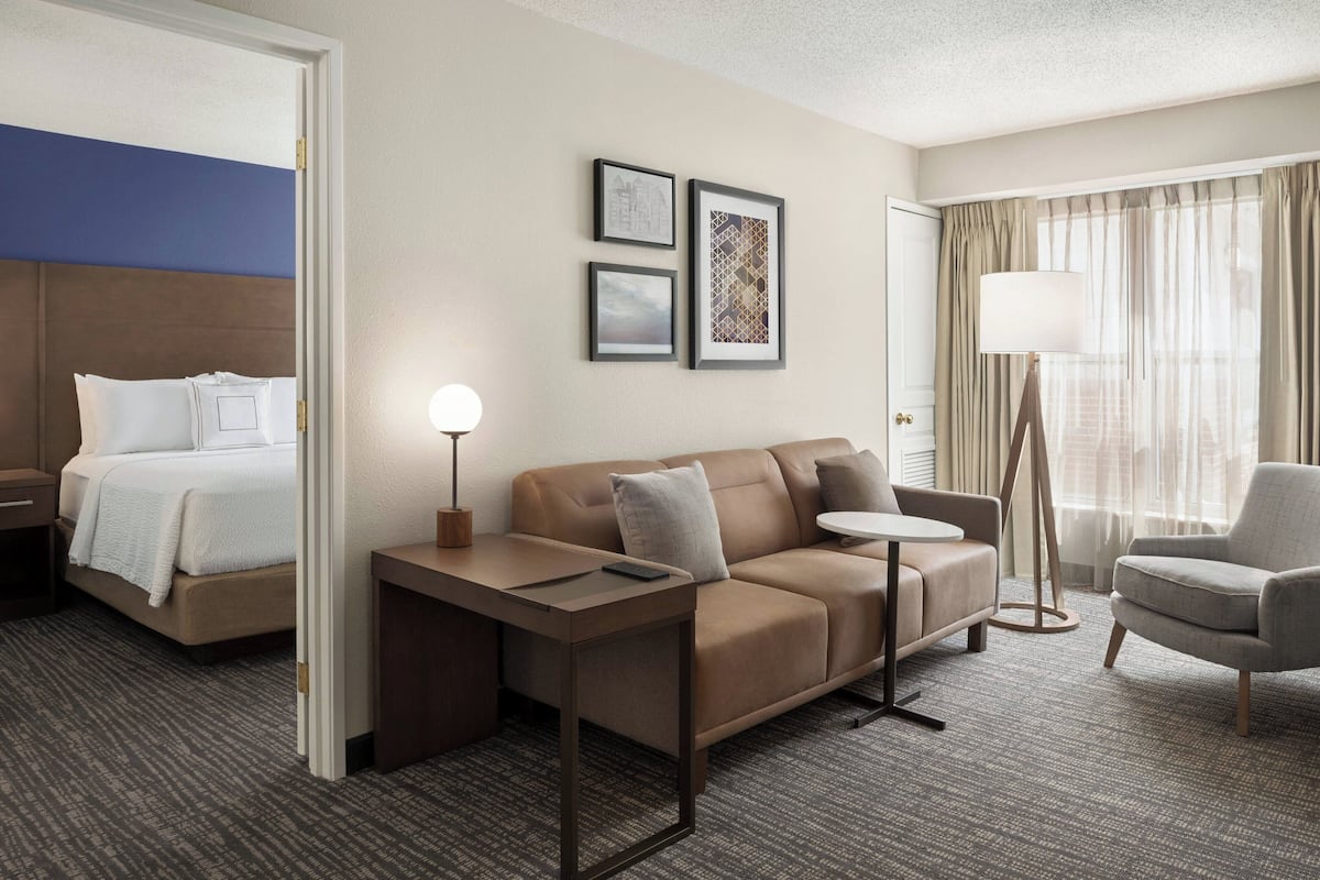 A hotel room with a bed, a sofa, a small table, a chair, and a lamp. There are framed pictures on the wall.