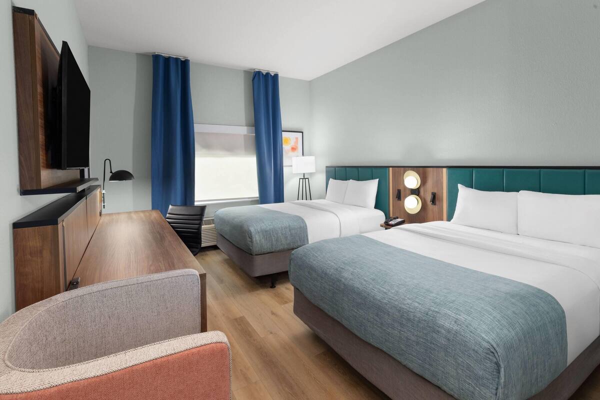 Ein Hotelzimmer mit zwei Betten, einem Schreibtisch, einem Stuhl, einem Fernseher und einem Fenster mit Vorhängen.