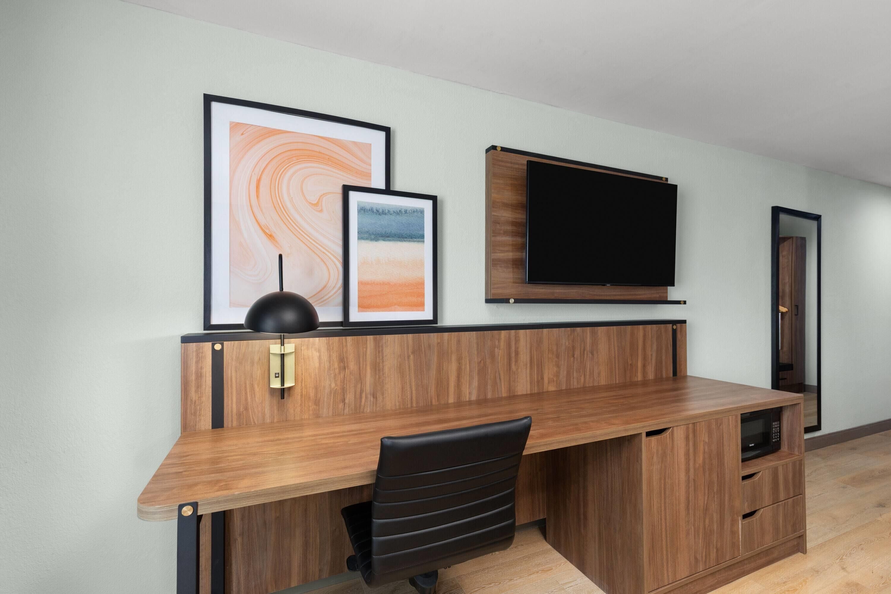 Ein moderner Arbeitsraum mit Schreibtisch aus Holz, schwarzem Bürostuhl, an der Wand montiertem Flachbildfernseher und gerahmten Kunstwerken.