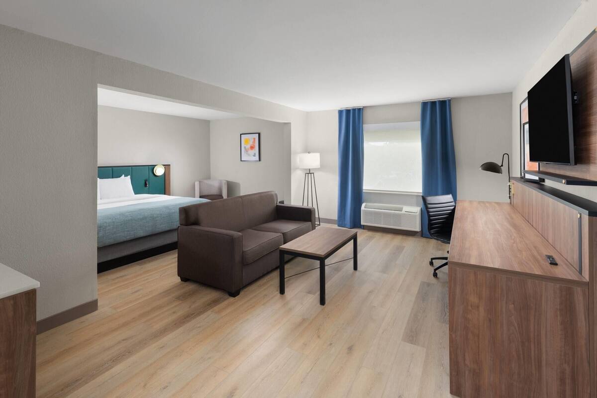 Ein modernes Hotelzimmer mit Bett, Sofa, Schreibtisch, Fernseher und einem großen Fenster mit Vorhängen.
