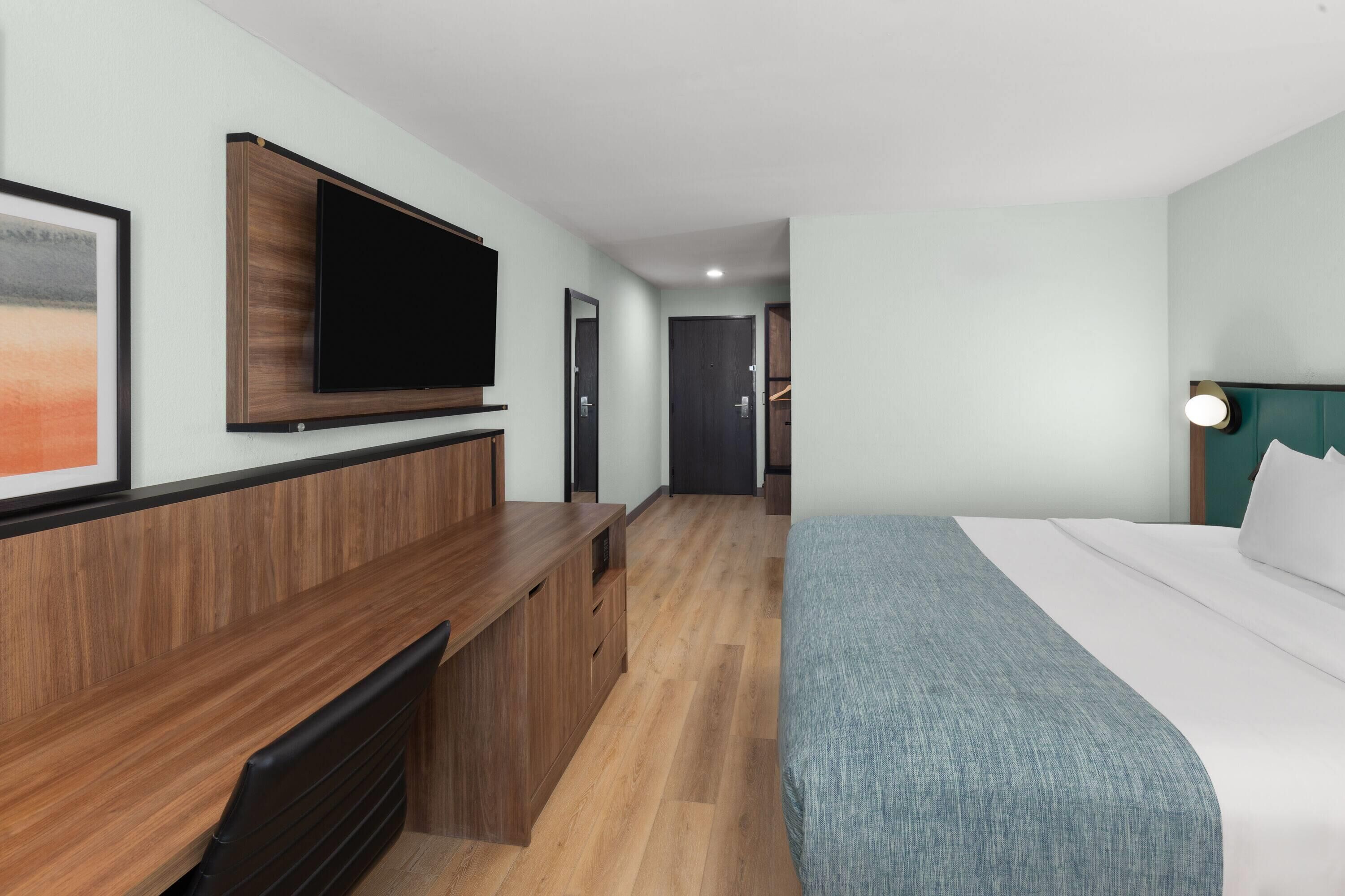 A hotel room with a bed, a desk with a chair, a flat-screen TV mounted on the wall, and a door leading to a closet.