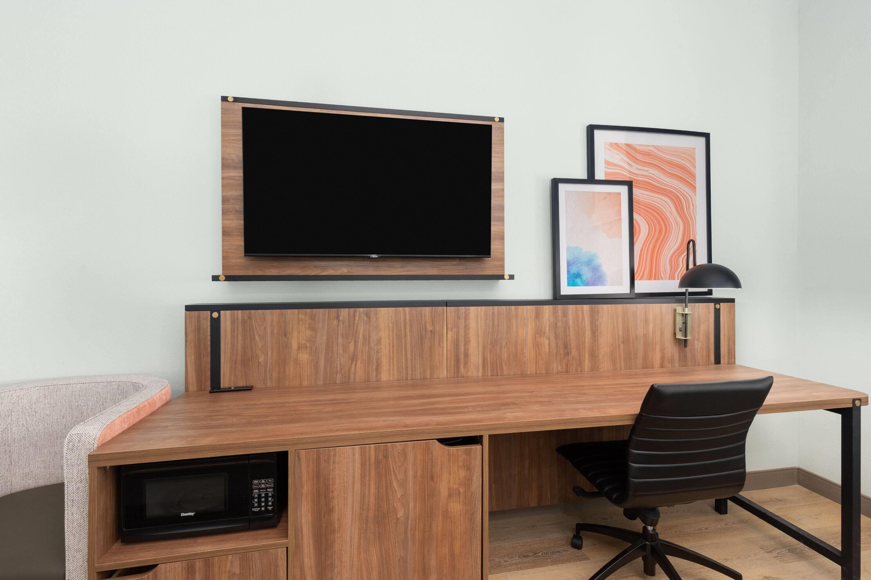Un espace de bureau moderne avec un bureau en bois, un téléviseur à écran plat fixé au mur, un fauteuil de bureau noir, un four à micro-ondes et des œuvres d’art encadrées accrochées au mur.