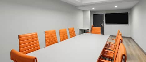 A conference room with a long table and orange chairs, a flat-screen TV, and a door.