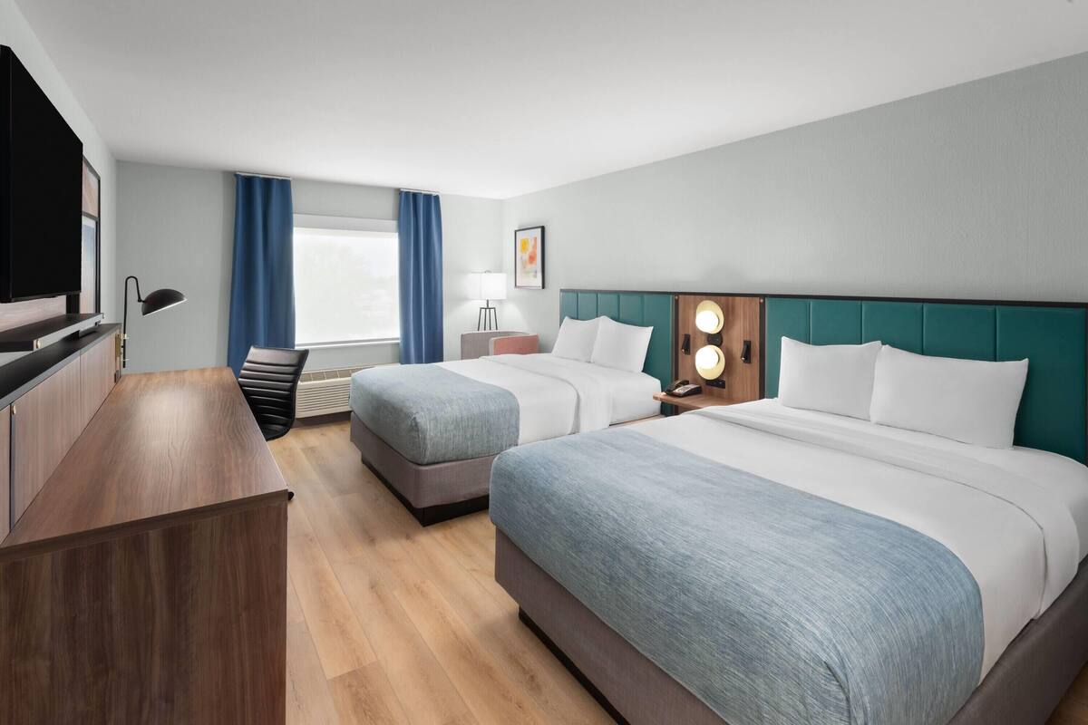 Ein Hotelzimmer mit zwei Betten, einem Schreibtisch, einem Fernseher und einem Fenster mit Vorhängen.