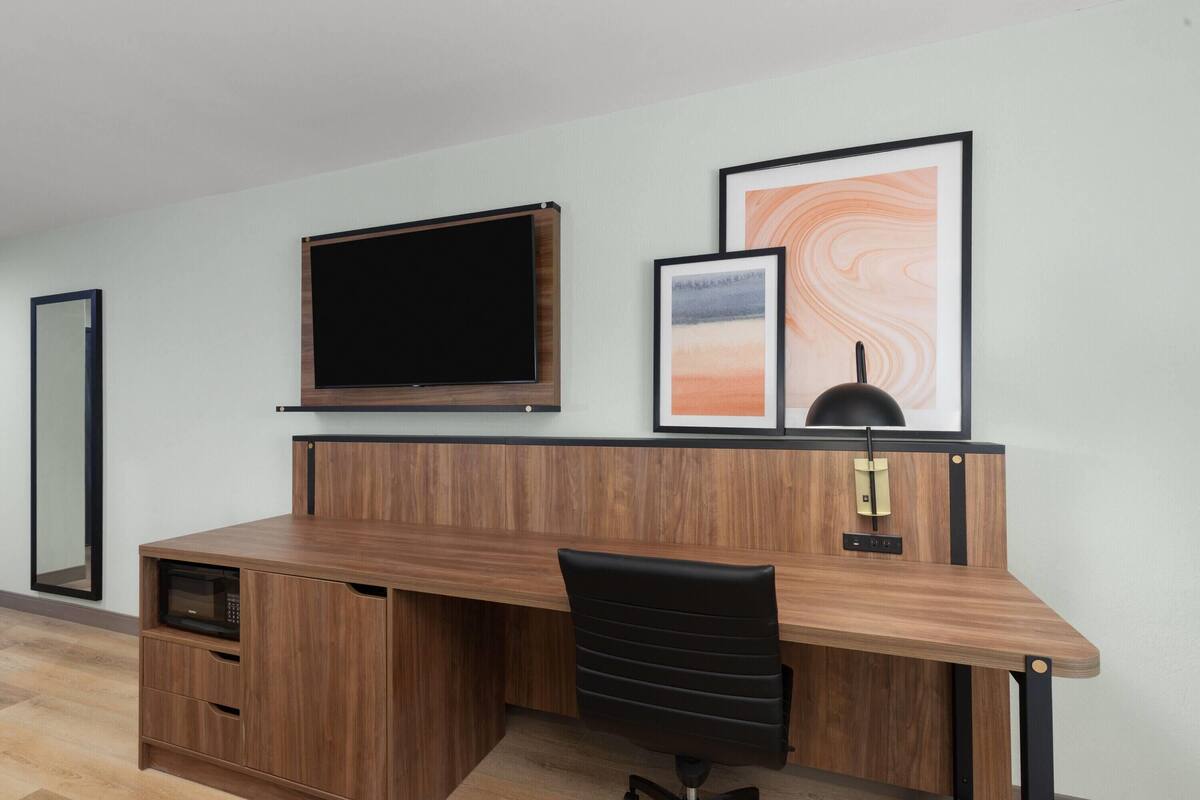 Ein moderner Arbeitsplatz mit einem Holztisch, einem schwarzen Bürostuhl, einem Wand-Fernseher und gerahmten Kunstwerken an der Wand.