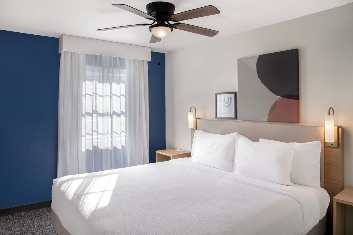 A hotel room with a large bed, a ceiling fan, a nightstand with a lamp, and a window with sheer curtains.