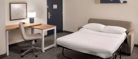 A hotel room with a bed, a desk with a chair, a lamp, and a painting on the wall.