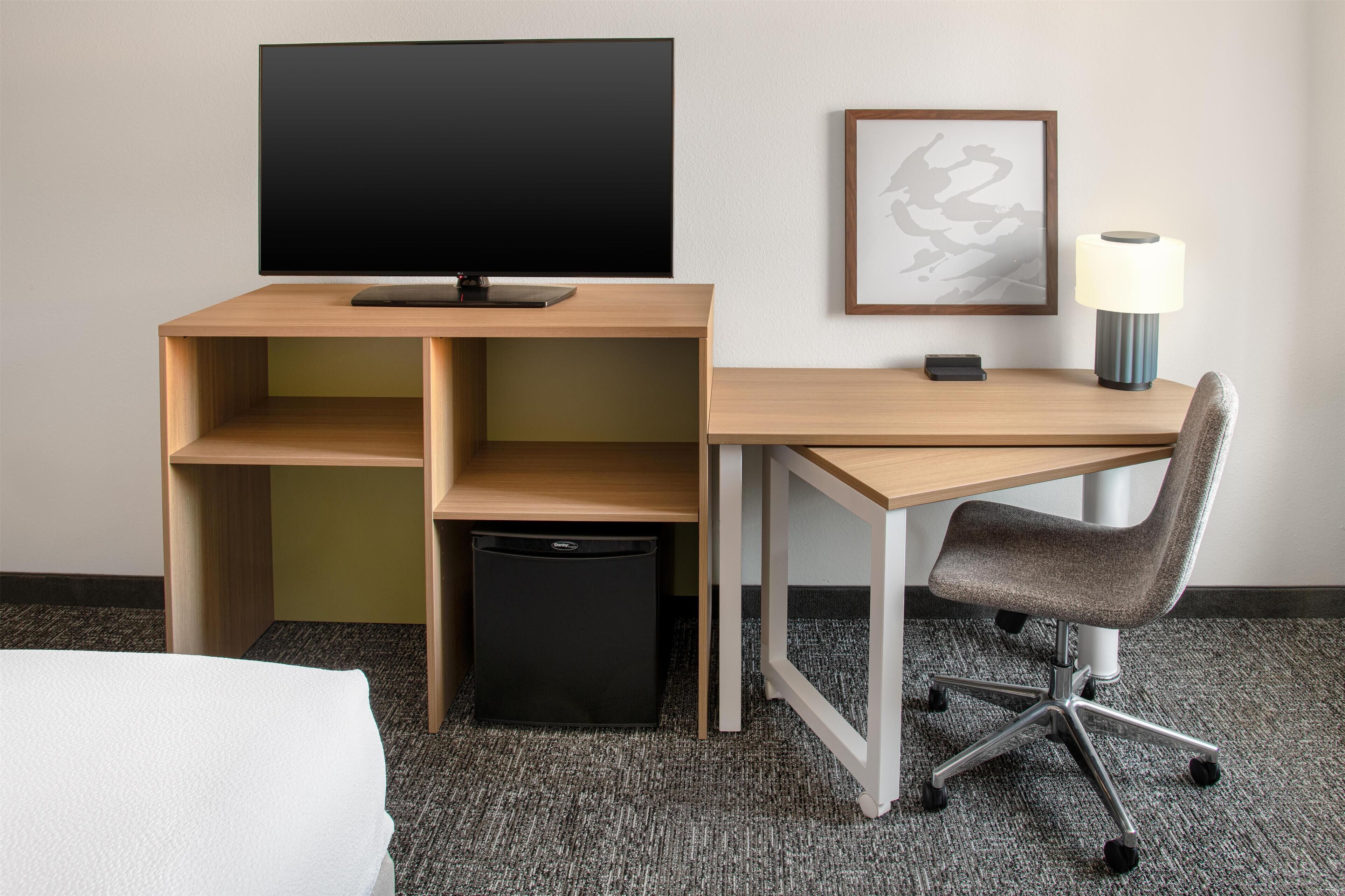 Een hotelkamer met een houten bureau, een televisie, een ingelijste foto, een bureaulamp, een stoel en een prullenbak.