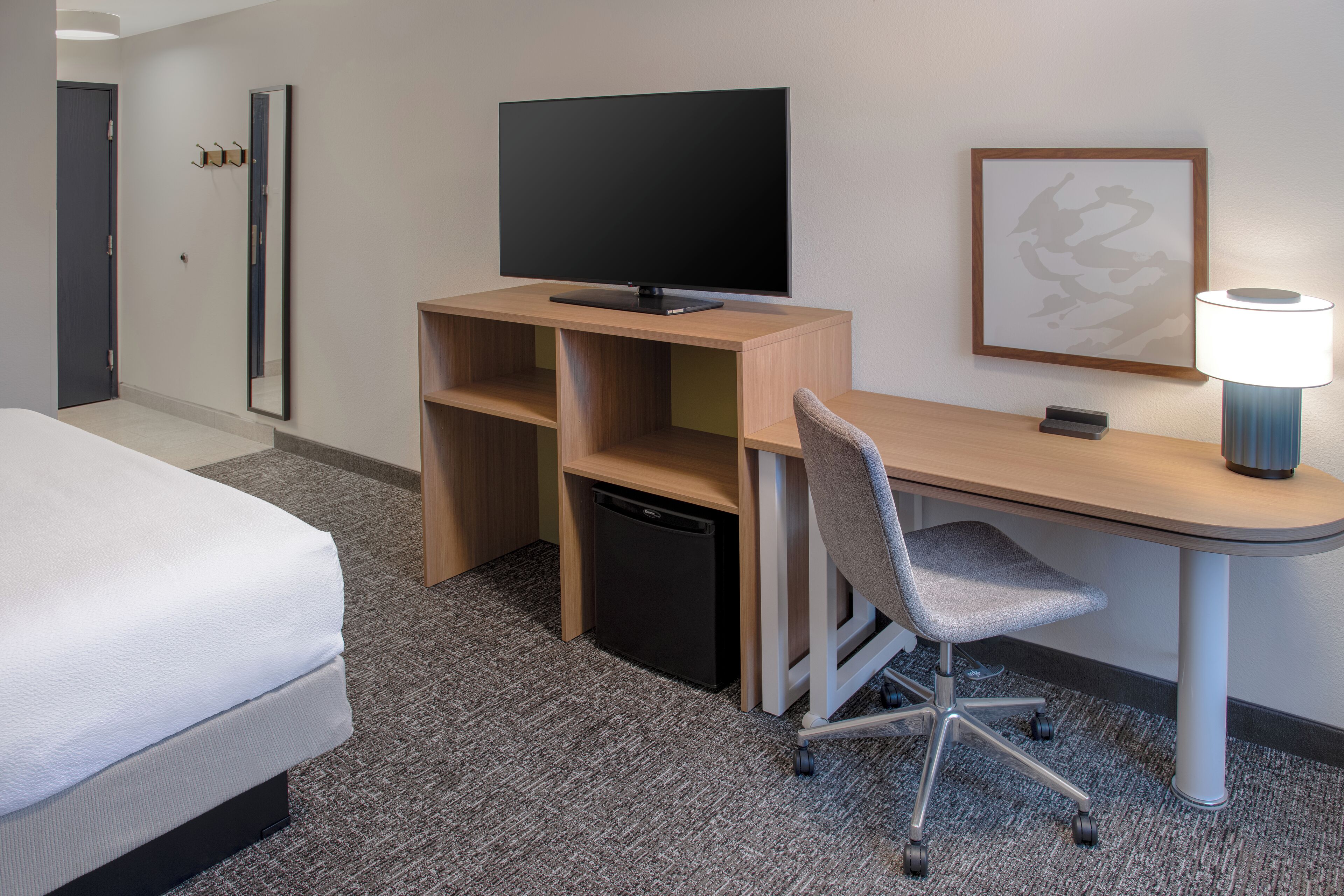 Een hotelkamer met een bed, een bureau met een televisie, een stoel, een lamp en een kleine koelkast.