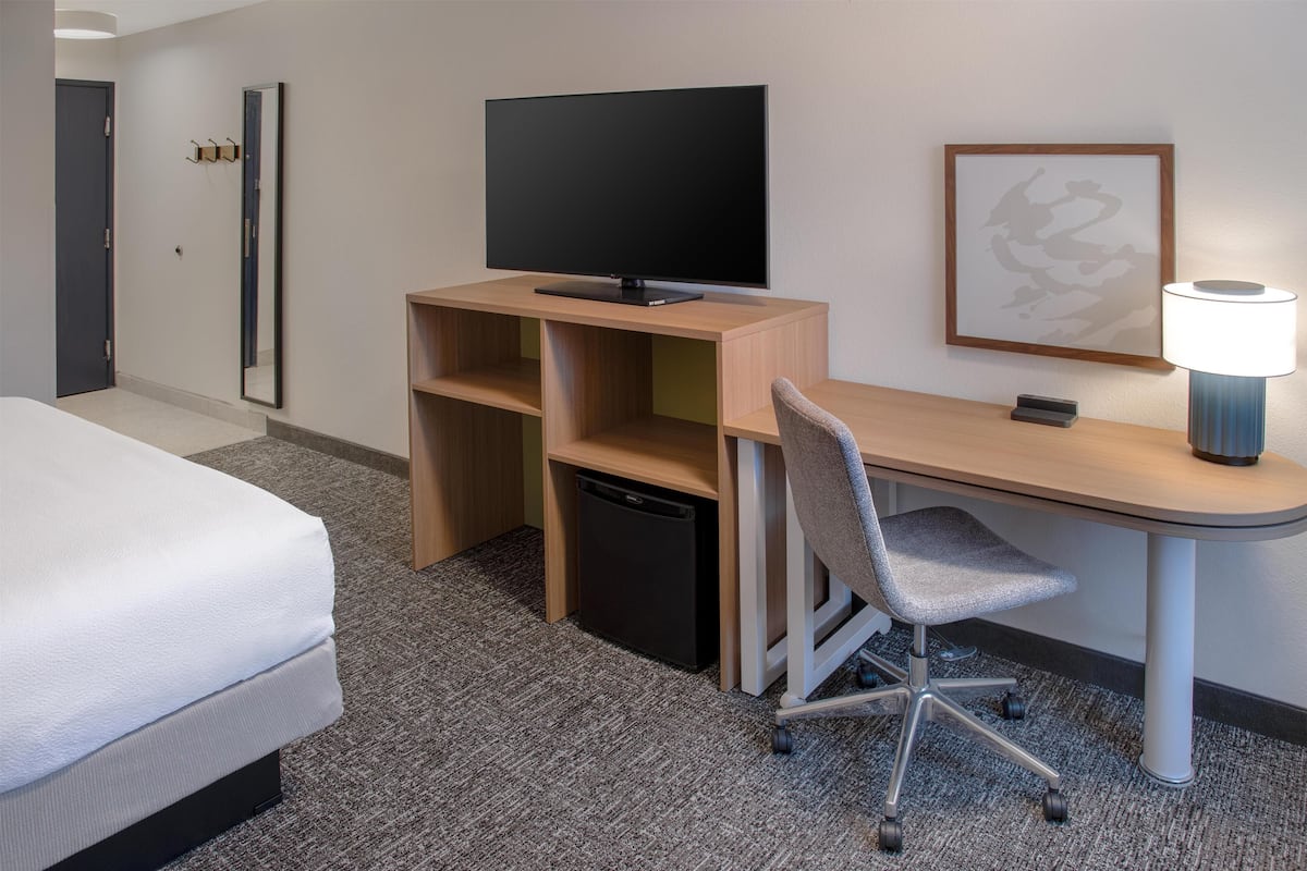 A hotel room with a bed, a desk with a television, a chair, a lamp, and a small refrigerator.