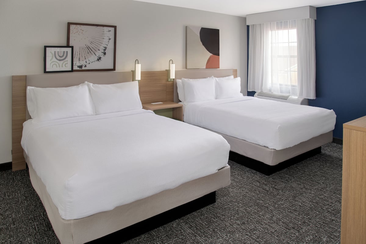 A hotel room with two beds, a nightstand, a lamp, and artwork on the wall.