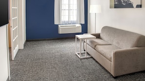 A room with a blue wall, a white air conditioning unit, a beige sofa, a small white side table, and a window with white curtains.