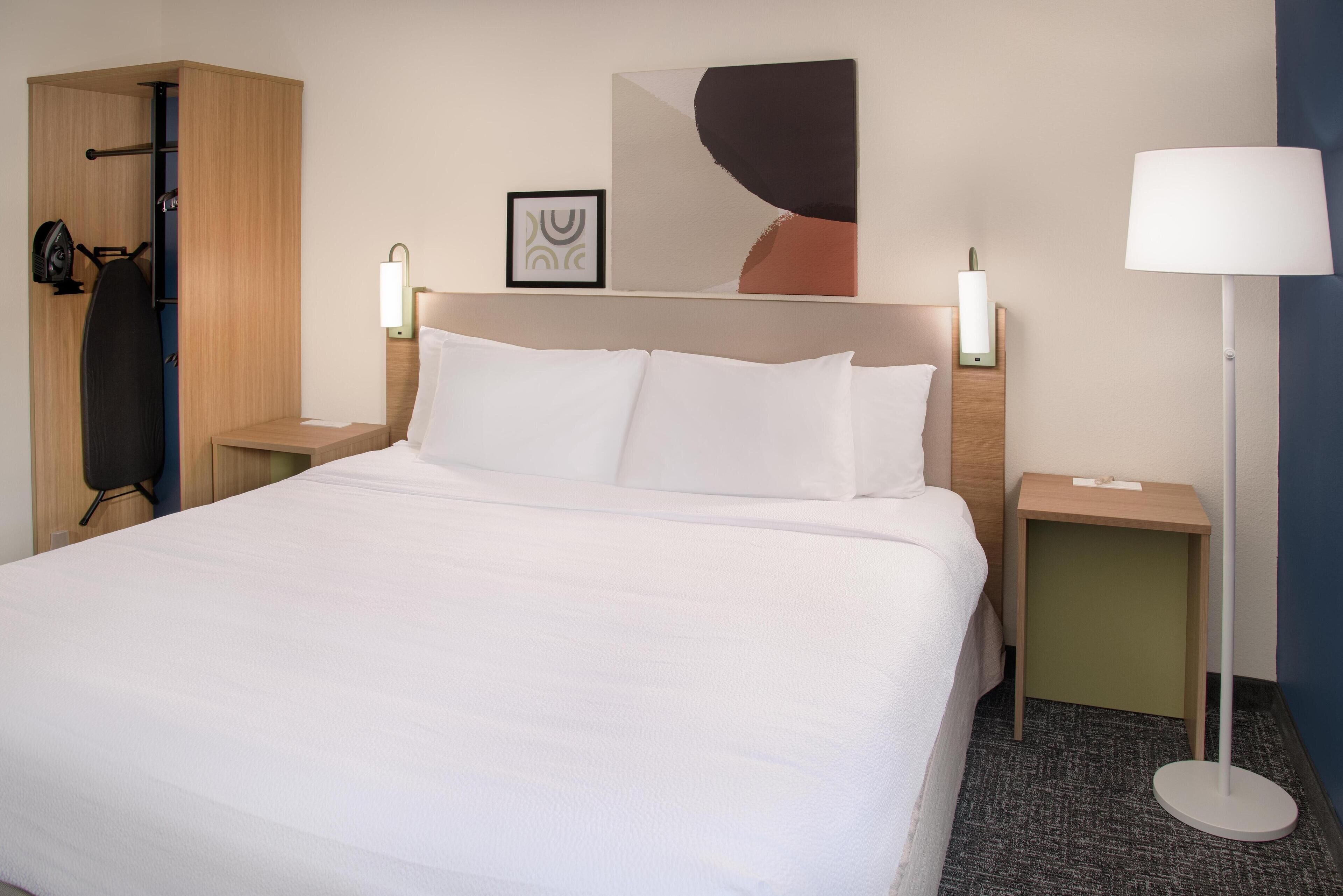 A hotel room with a large bed, two bedside tables, a standing lamp, a wardrobe, and a wall art piece.