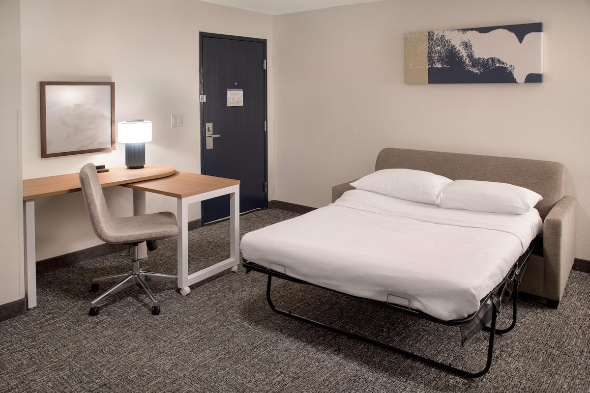 A hotel room with a bed, a desk, a chair, a lamp, and a door.