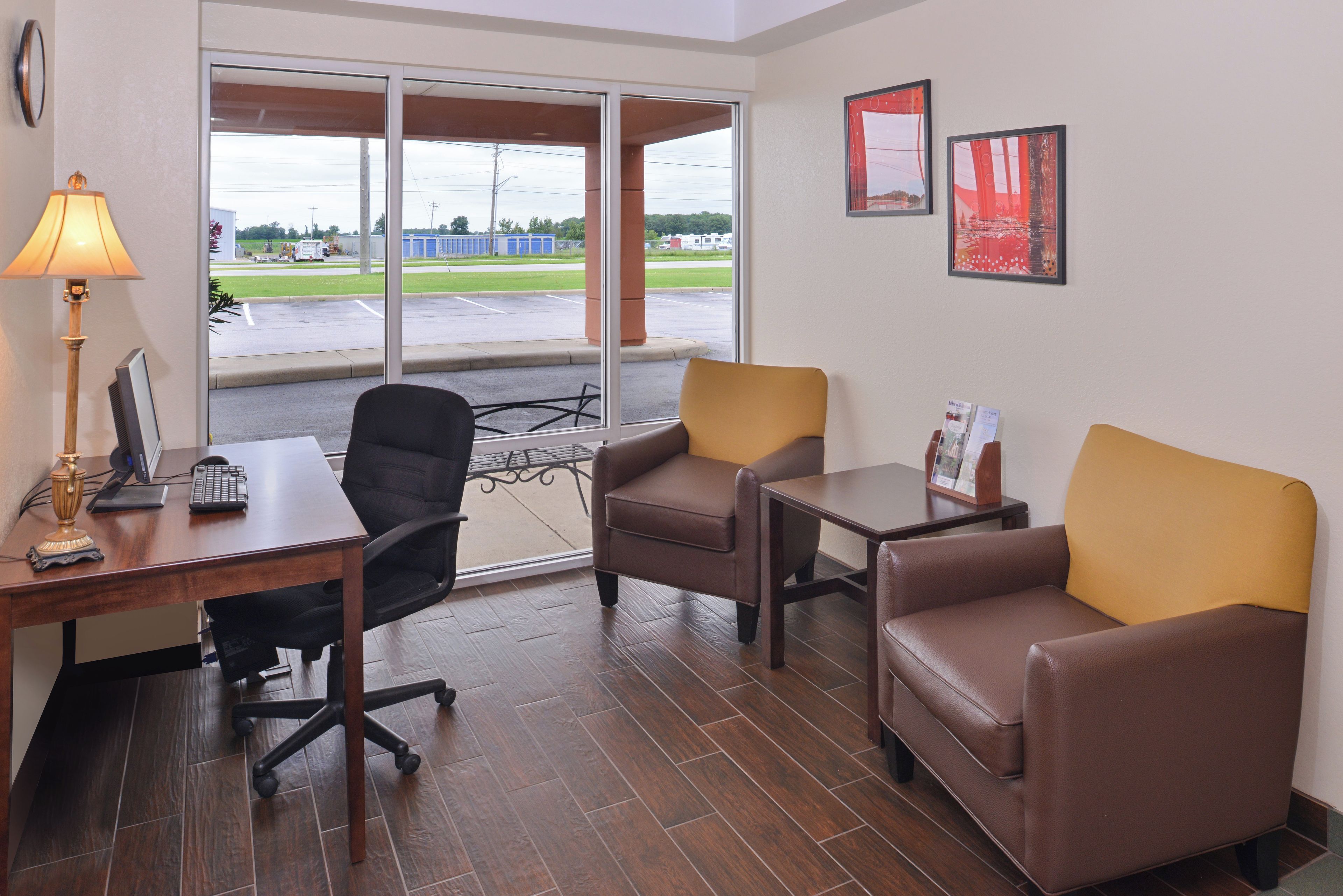 Habitación de hotel con escritorio, dos sillas, una lámpara y vista a un estacionamiento desde la puerta.