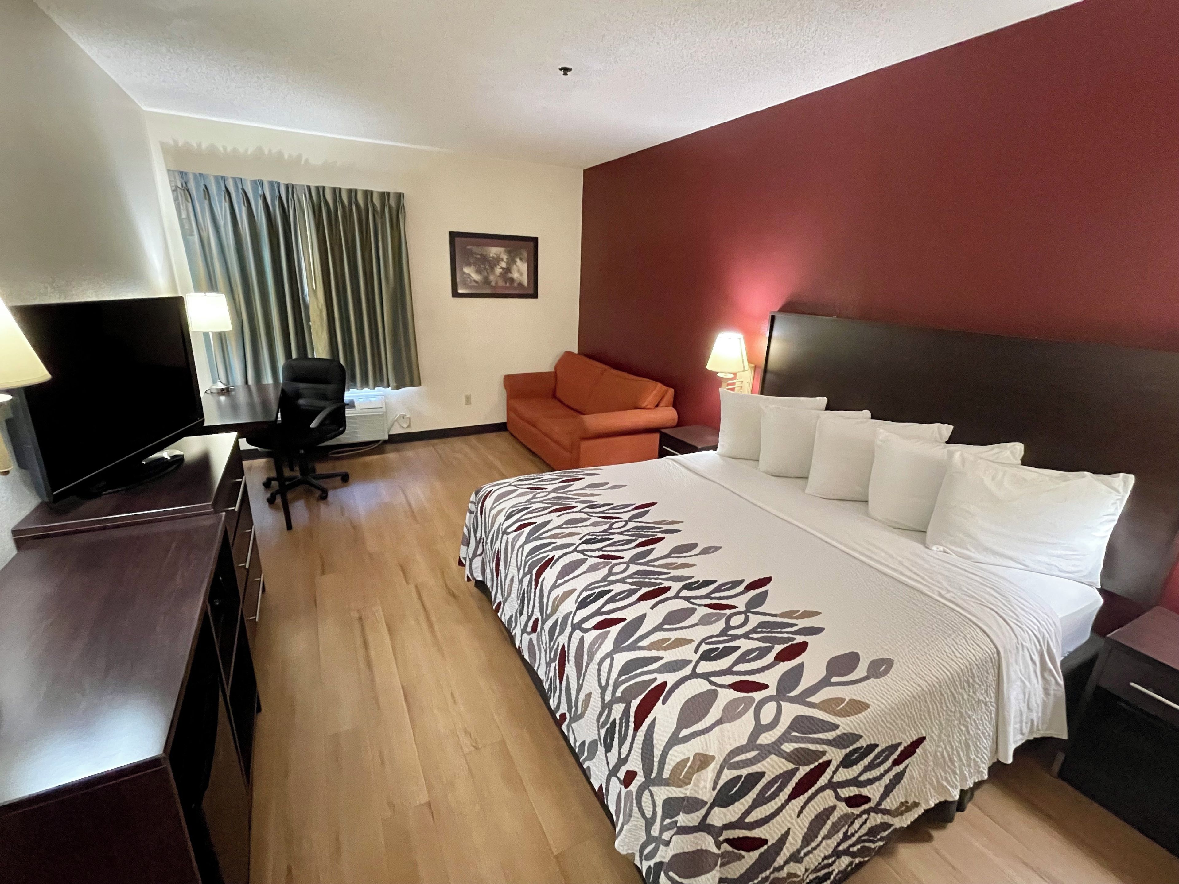 Habitación de hotel con una cama, un escritorio con computadora, una silla, un televisor y una ventana con cortinas.