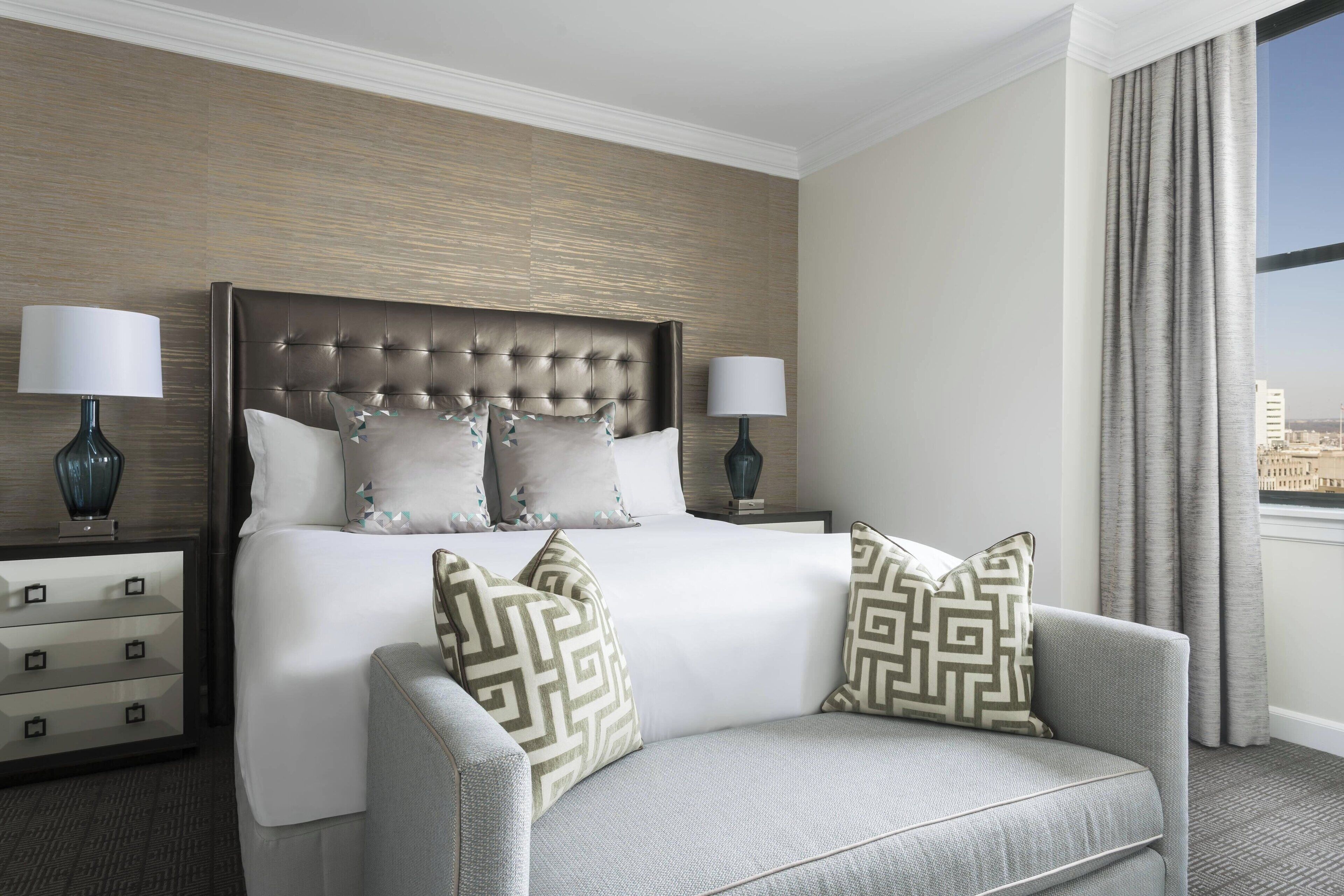 A hotel room with a large bed, two bedside tables with lamps, a grey sofa, and a view of the city through the window.