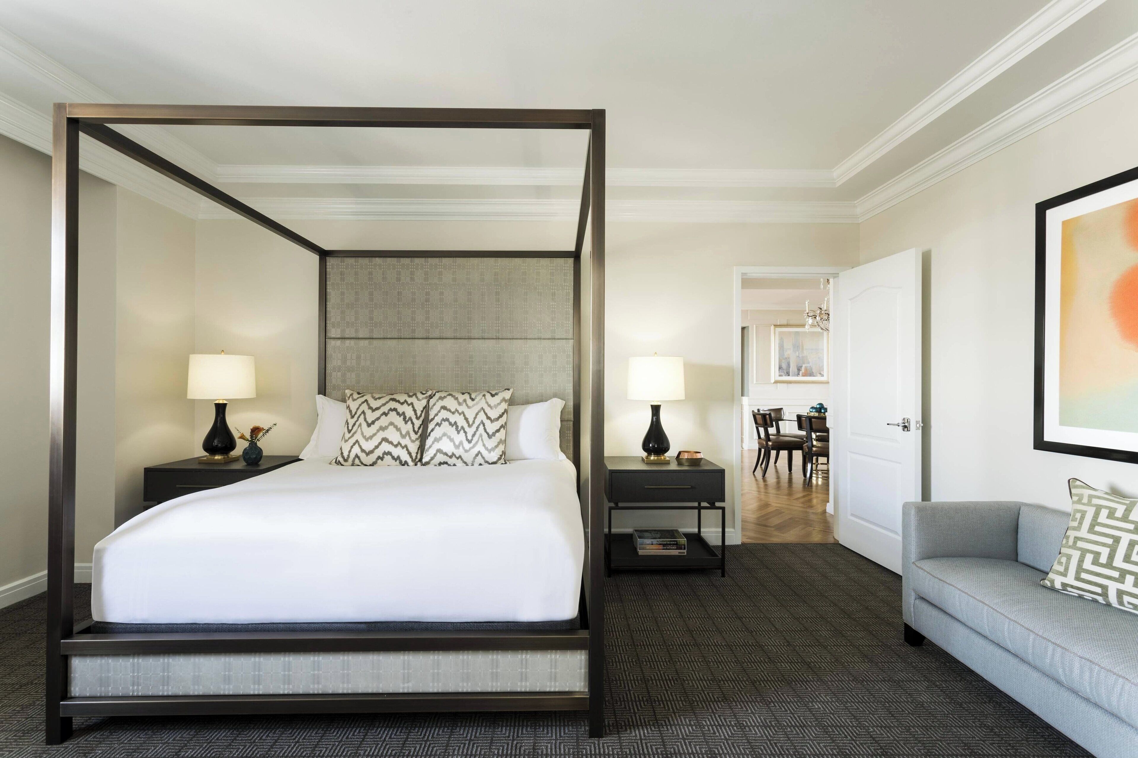 A four-poster bed with a grey upholstered headboard, two bedside tables with lamps, a grey sofa with patterned cushions, and a framed abstract painting on the wall.