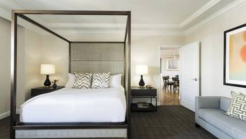 A four-poster bed with a grey upholstered headboard, two bedside tables with lamps, a grey sofa with patterned cushions, and a framed abstract painting on the wall.