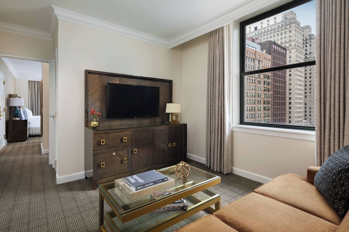 Habitación de hotel con un televisor de pantalla plana grande montado en un mueble de madera oscura, una mesa de centro de vidrio con libros y objetos decorativos, un sofá cómodo y vistas a edificios de la ciudad a través de una ventana grande con cortinas.