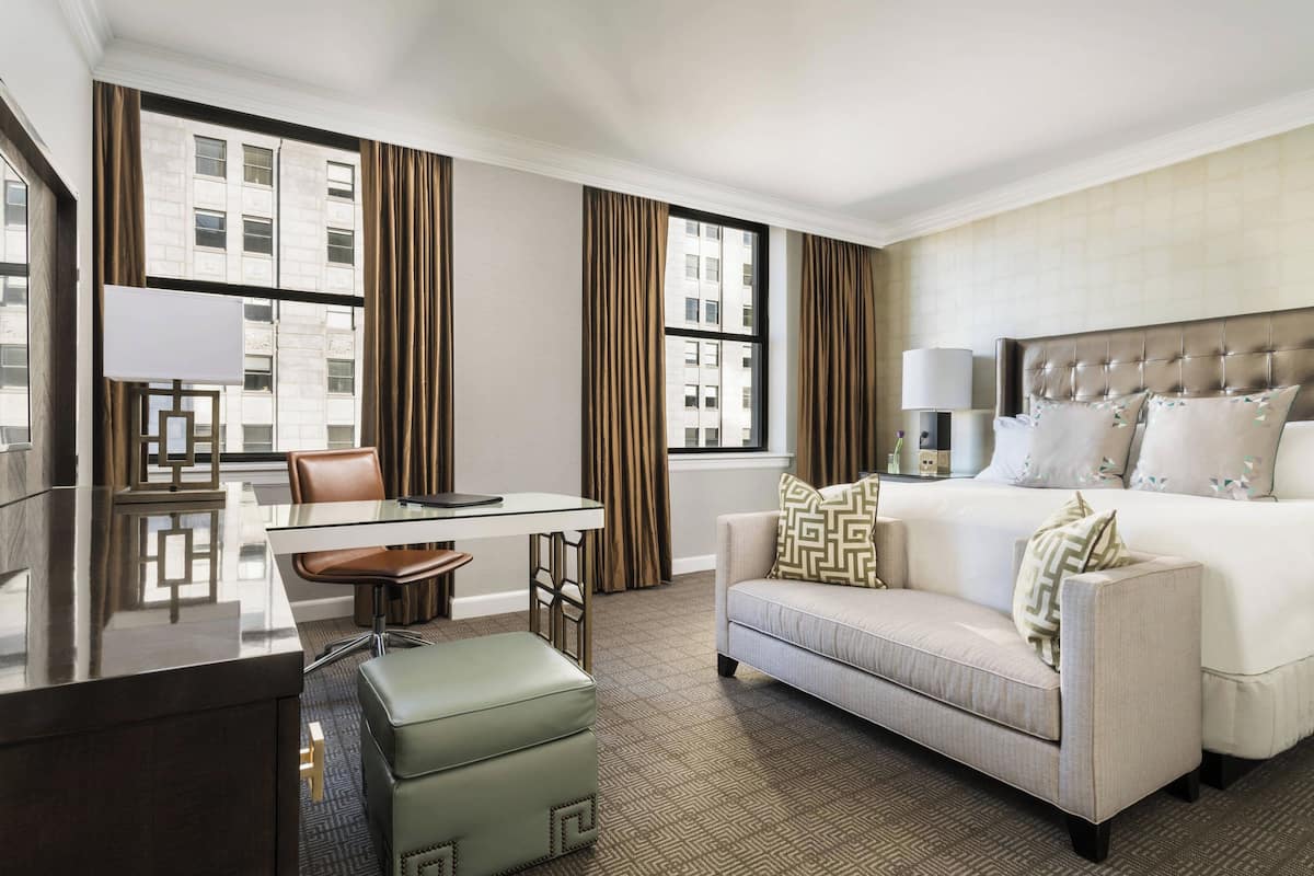 Habitación de hotel con cama, sofá, mesa de comedor con sillas y vistas a un edificio a través de amplios ventanales.