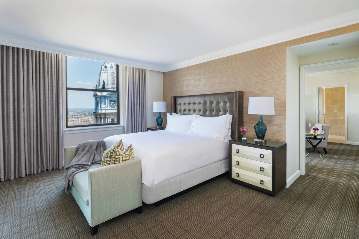 A hotel room with a large bed, a nightstand, a chair, a desk, and a view of a cityscape.