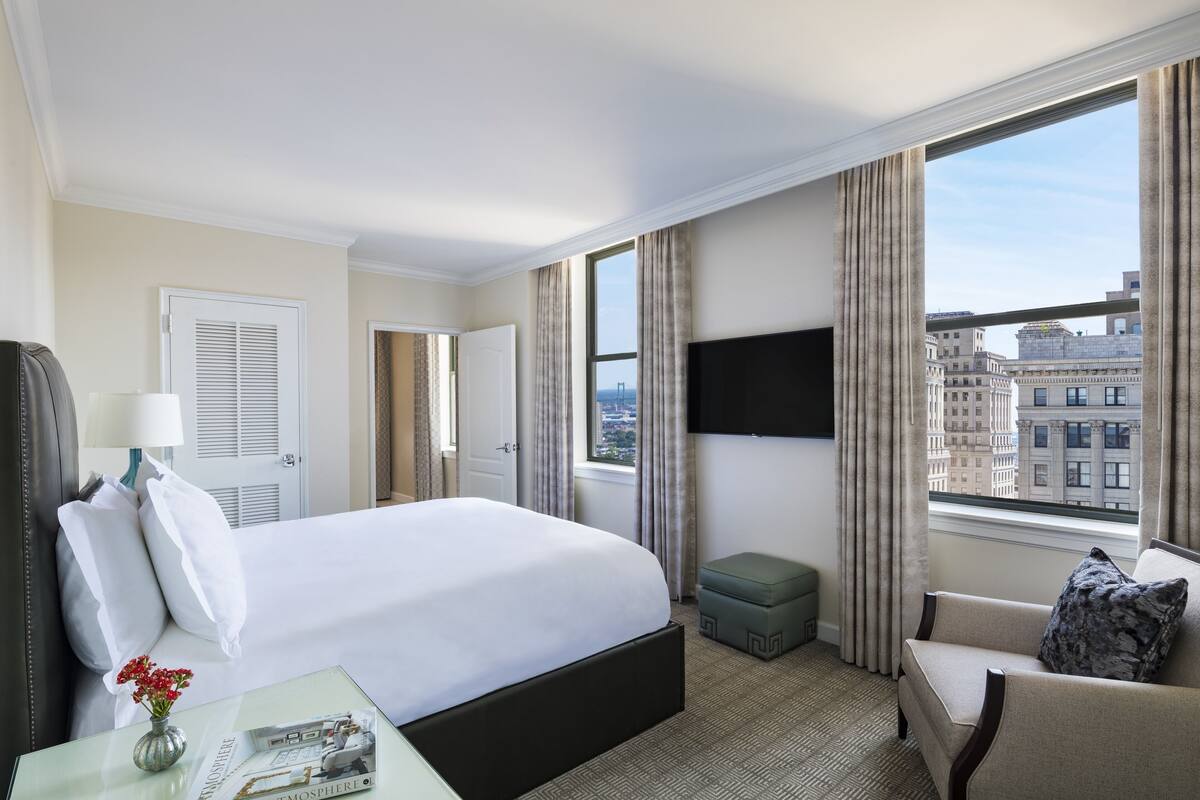 A hotel room with a large bed, a TV, a sofa, and a window with a city view.