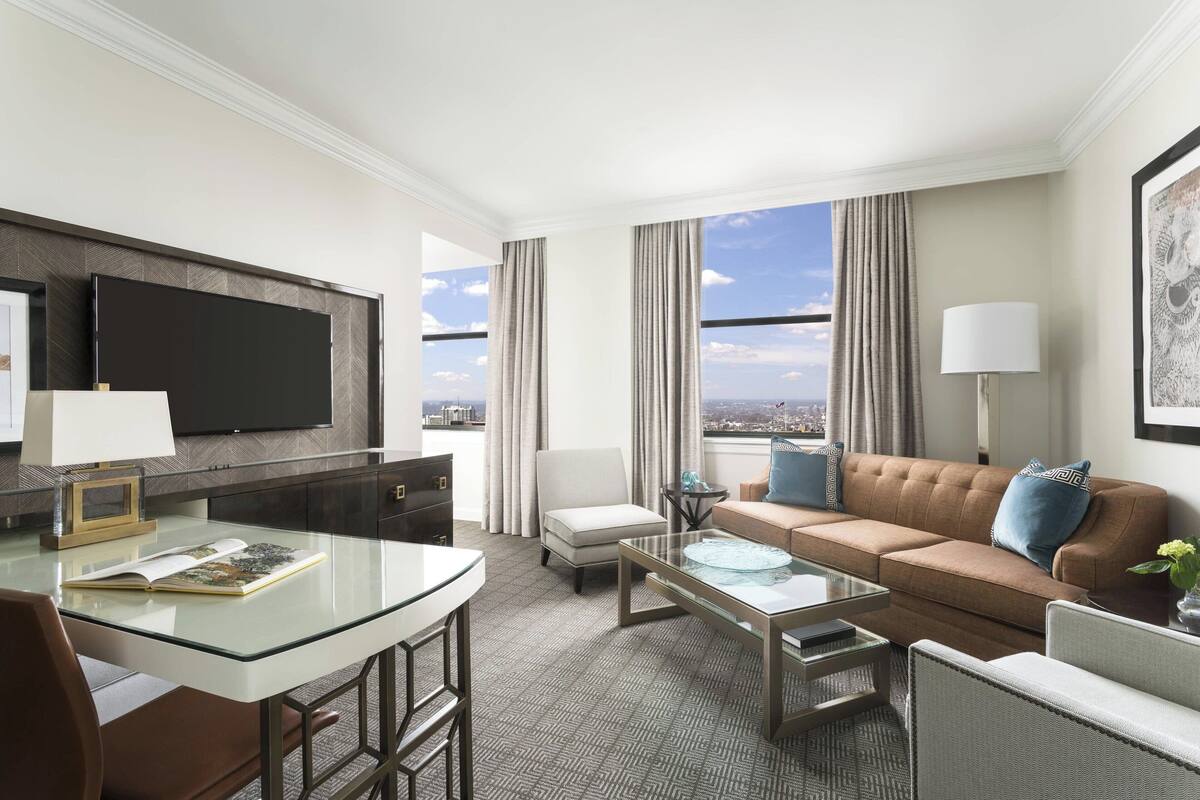 A modern hotel room with a glass dining table, a sofa, a TV mounted on the wall, and a view of the city.