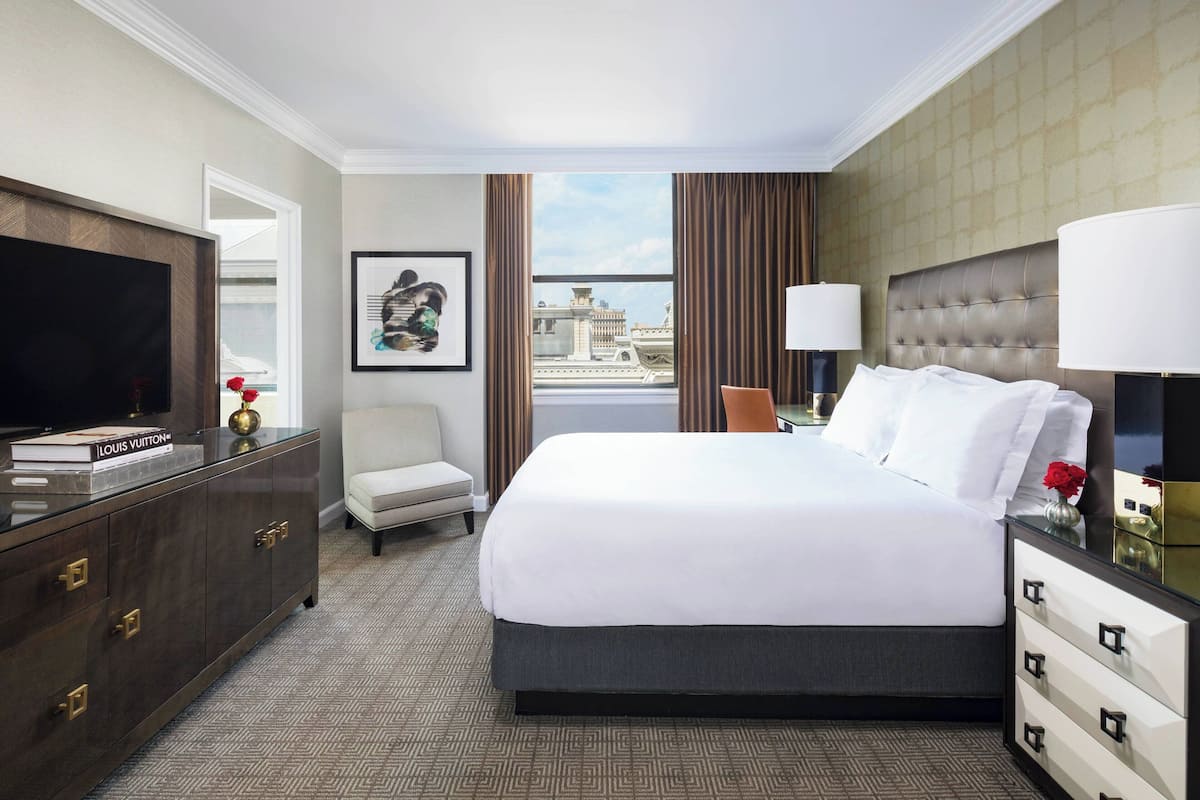 Una habitación de hotel con una cama grande, televisión, escritorio con lámpara, silla y vista a la ciudad.