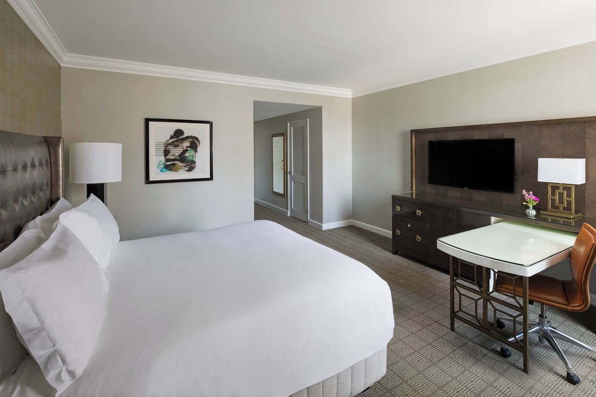 Una habitación de hotel con una cama grande, un escritorio con silla, un televisor de pantalla plana, un aparador y un cuadro en la pared.