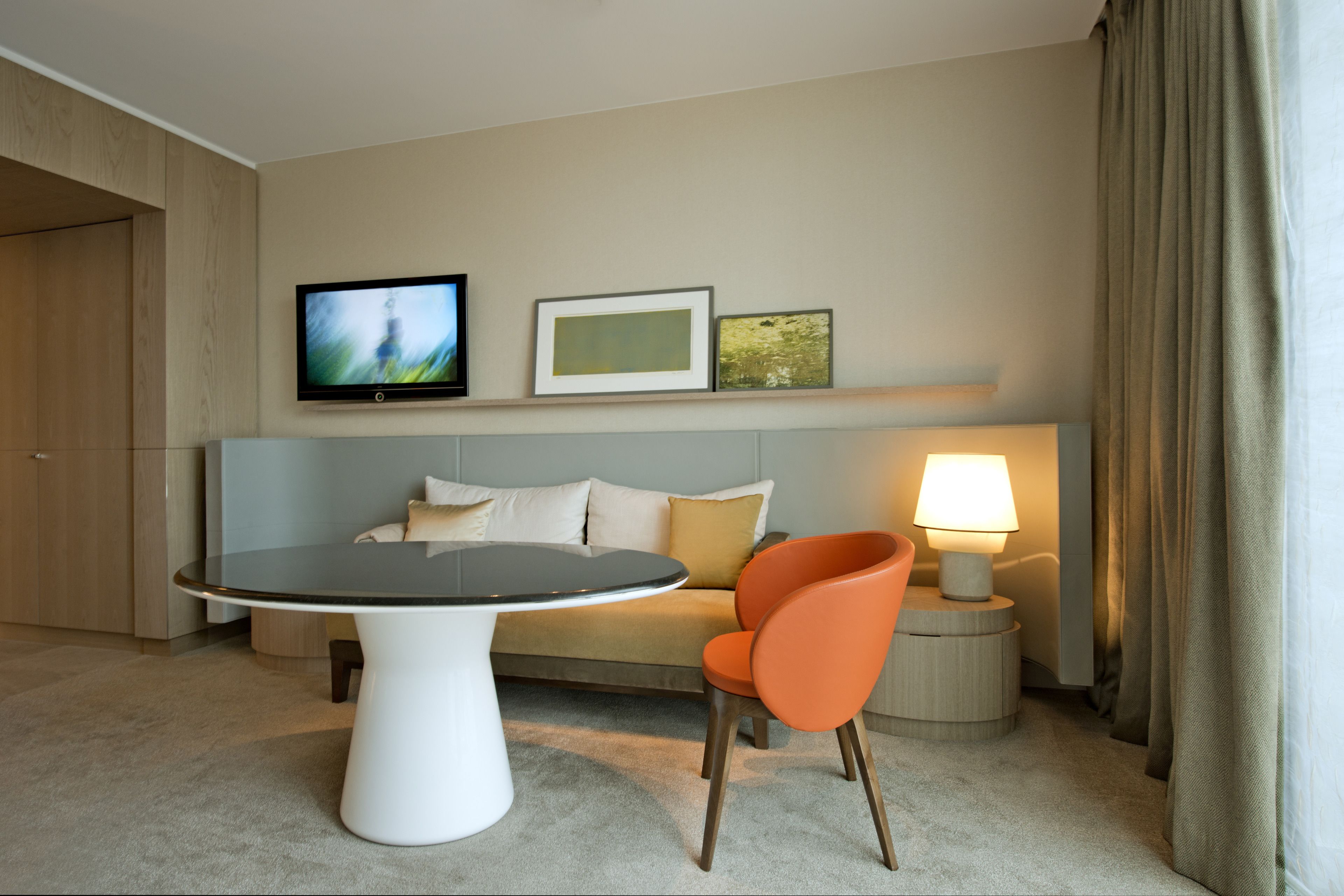 Quarto de hotel com uma mesa redonda, um sofá, uma poltrona laranja, uma TV fixada na parede e um abajur em uma mesinha de cabeceira.