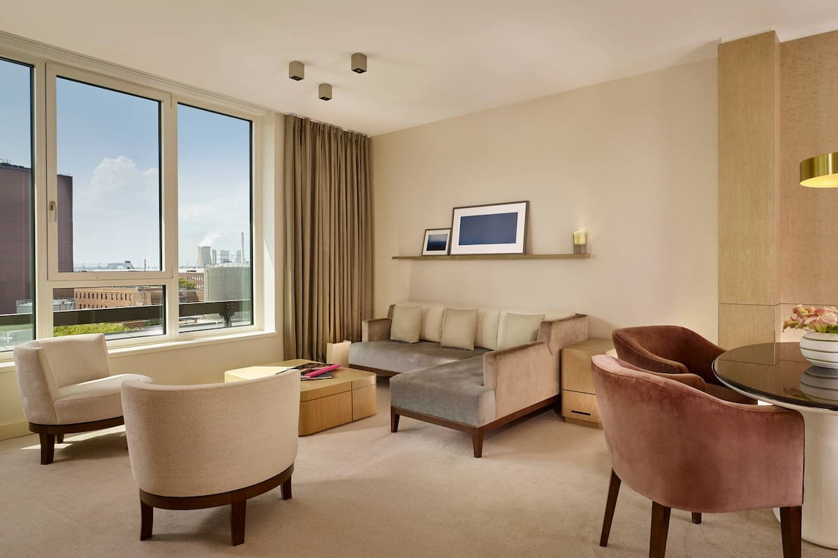 Ein modernes Hotelzimmer mit großem Fenster, einer Couch, zwei Sesseln, einem kleinen Tisch und einem Essbereich.