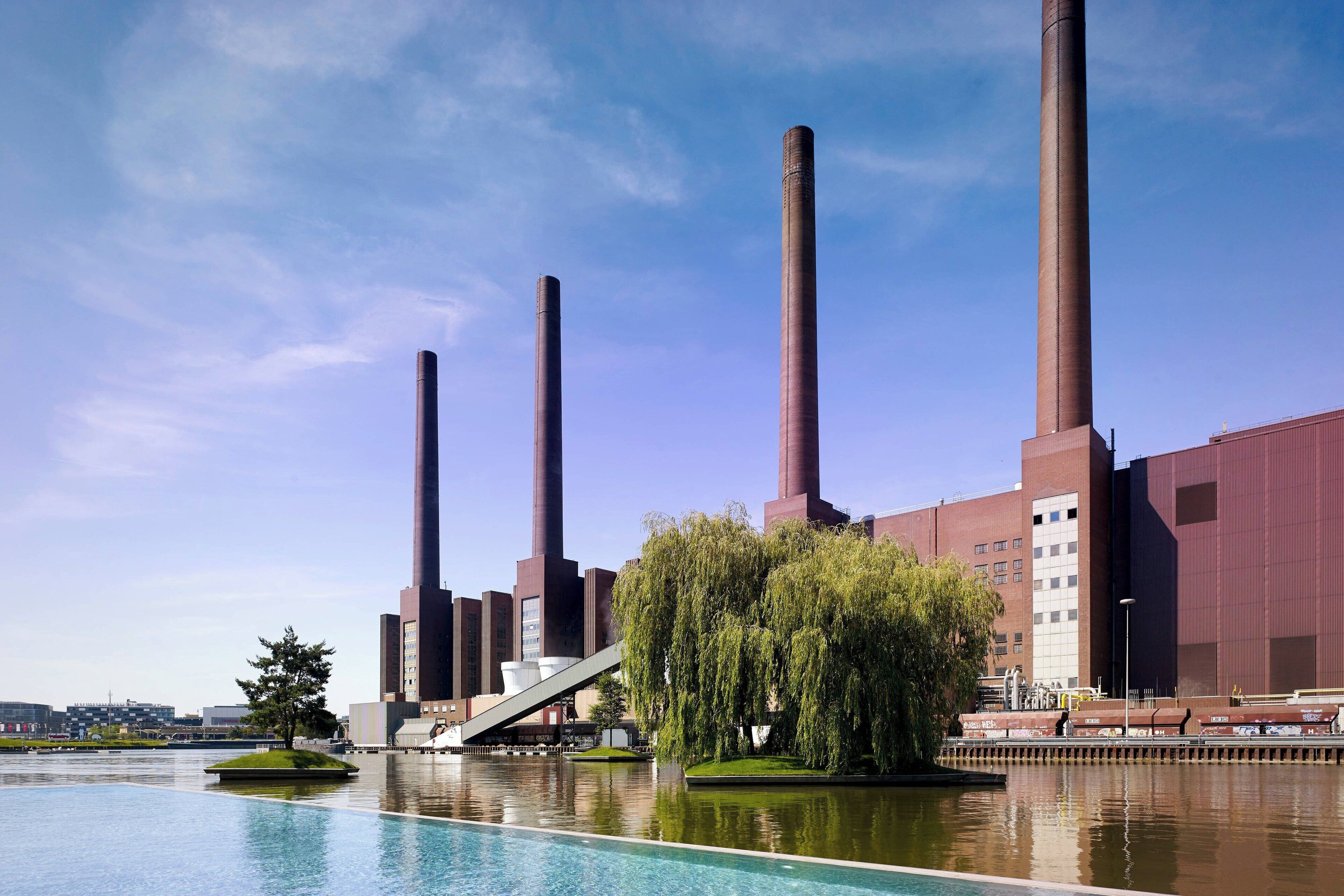 Een groot industriële complex met hoge schoorstenen en een rustig waterlichaam op de voorgrond.