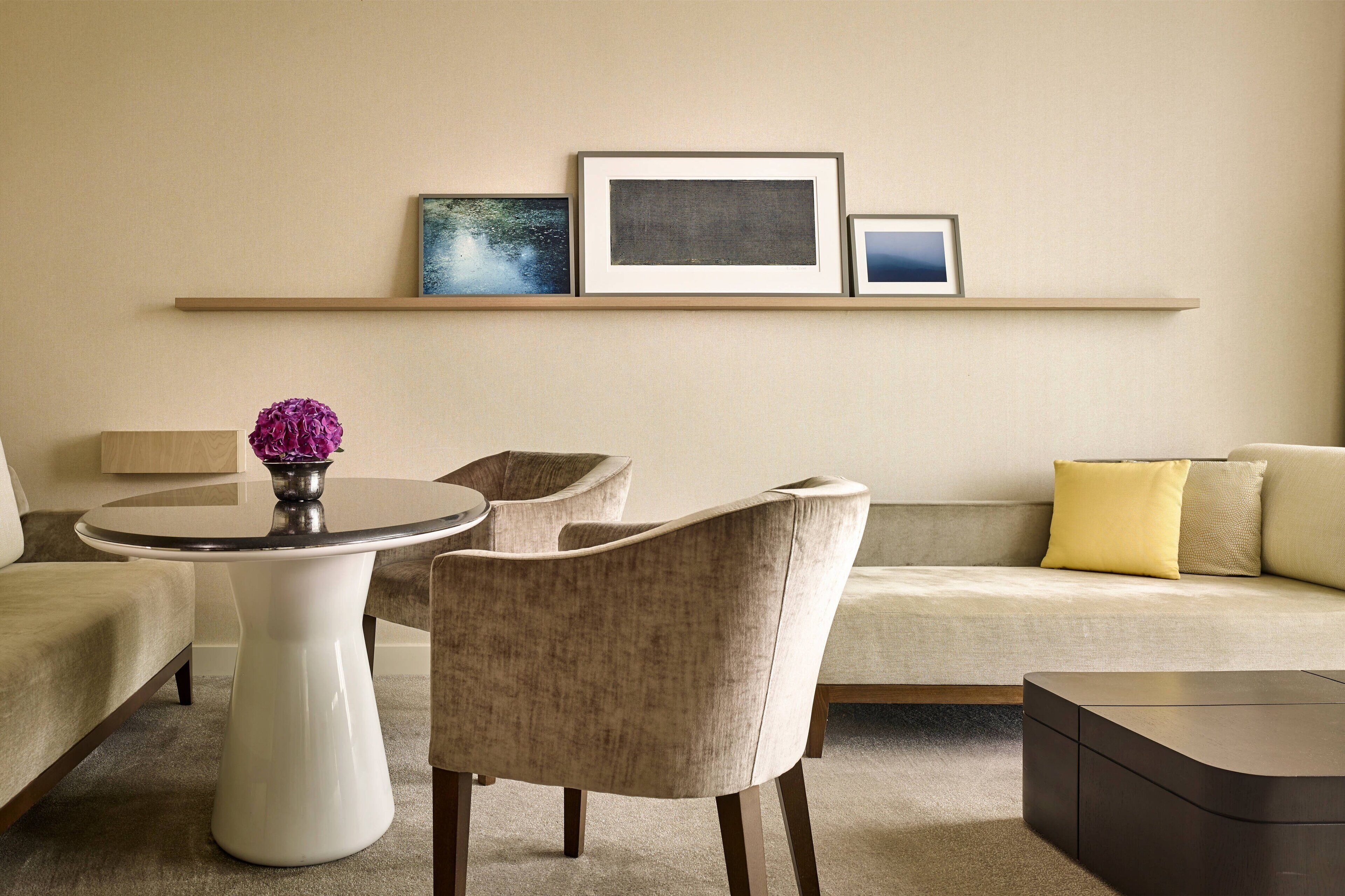 A modern living room with a round white table, two beige armchairs, a beige sofa with yellow and beige cushions, and a dark brown coffee table. There are three framed pictures on a wall-mounted shelf above the sofa.