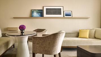 A modern living room with a round white table, two beige armchairs, a beige sofa with yellow and beige cushions, and a dark brown coffee table. There are three framed pictures on a wall-mounted shelf above the sofa.