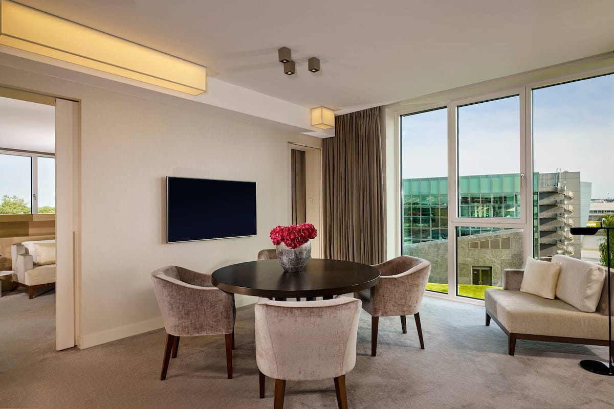 Ein modernes Hotelzimmer mit einem runden Esstisch, vier Stühlen, einem Flachbildfernseher und einem großen Fenster mit Blick auf ein Gebäude.