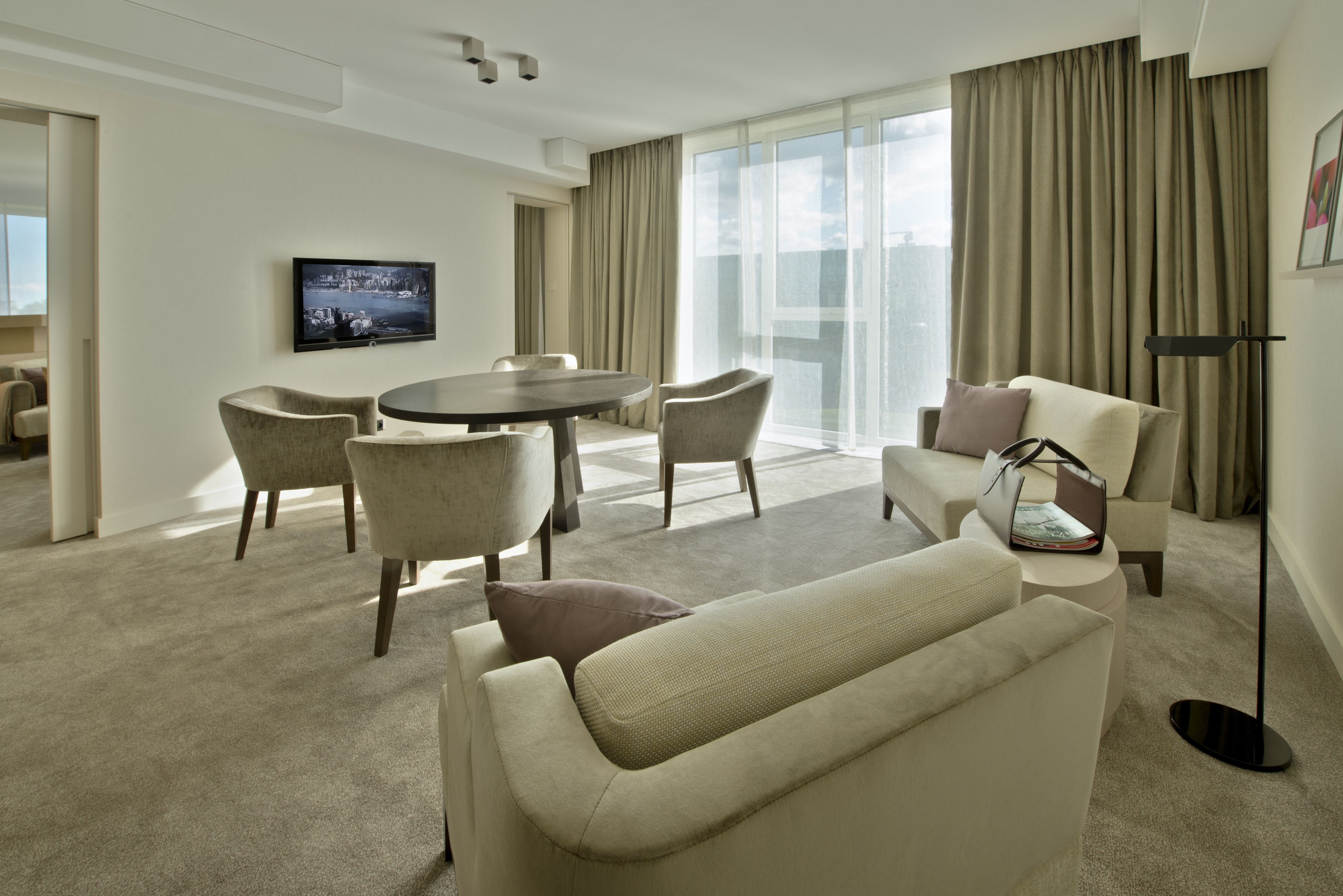 Ein modernes Hotelzimmer mit einem runden Esstisch, gemütlicher Sitzgelegenheit, einem Flachbildfernseher und großen Fenstern mit Vorhängen.