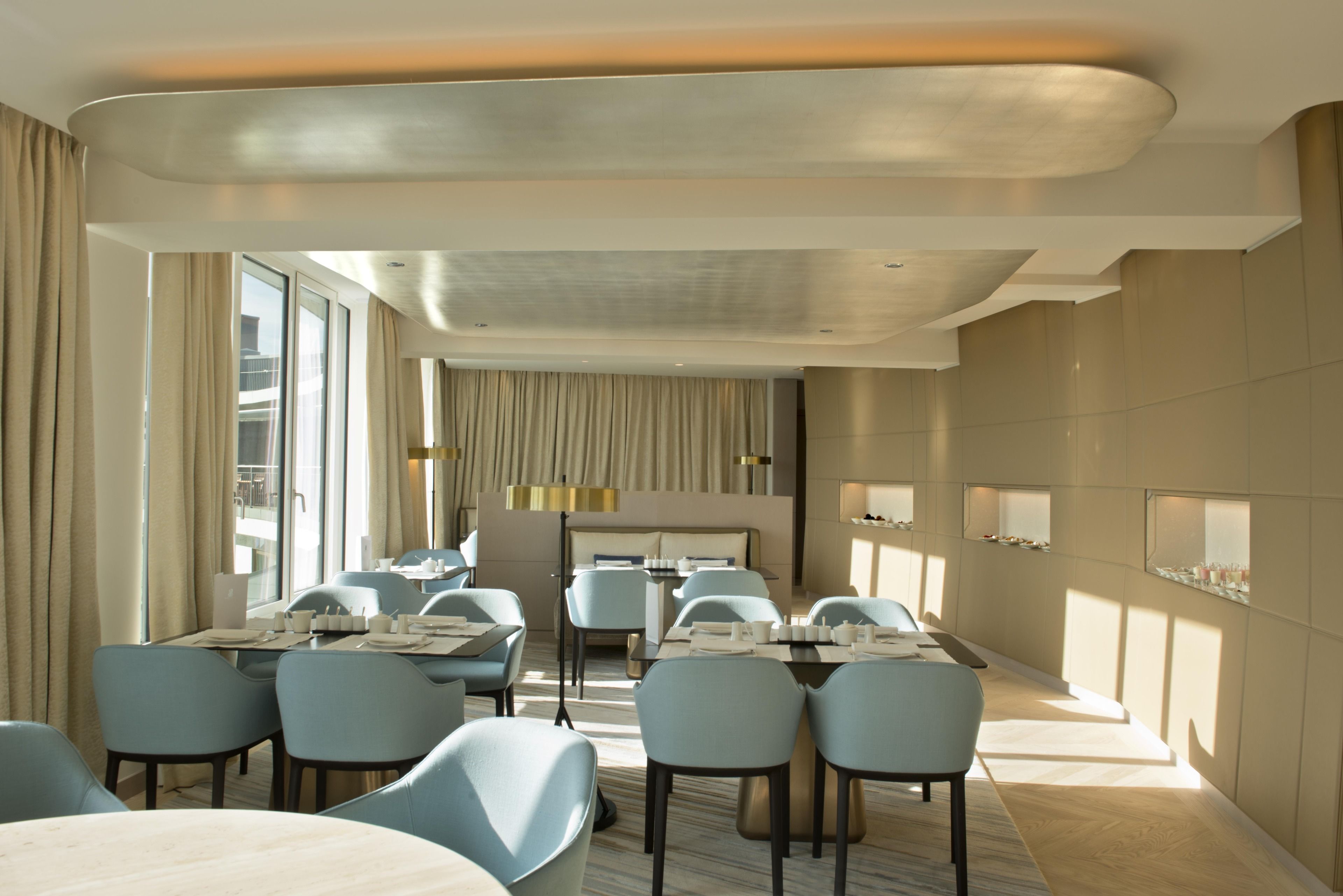 A modern hotel dining area with multiple tables and chairs, large windows, and a contemporary interior design.
