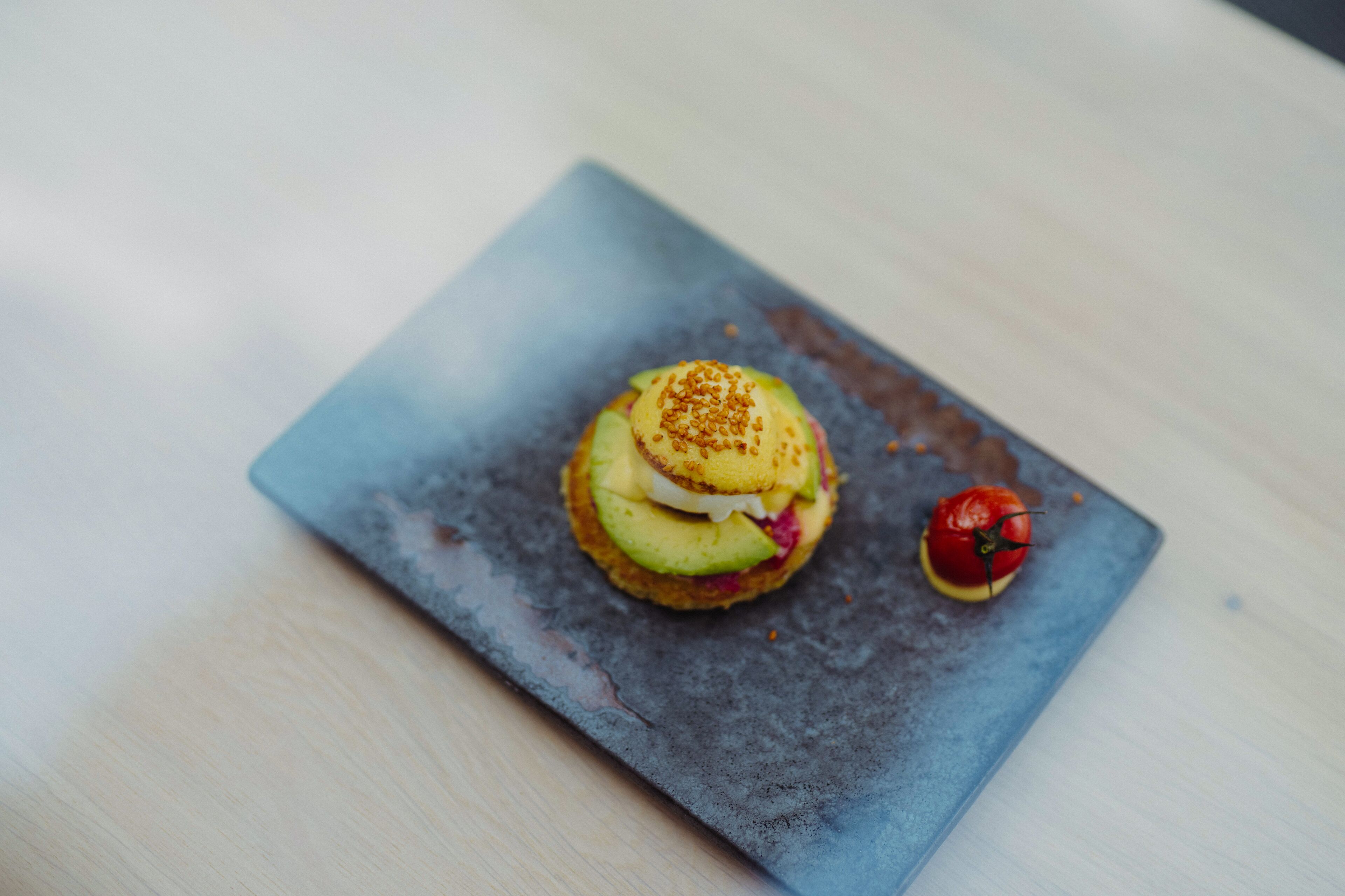 A square plate with a sandwich and a tomato on a wooden table.