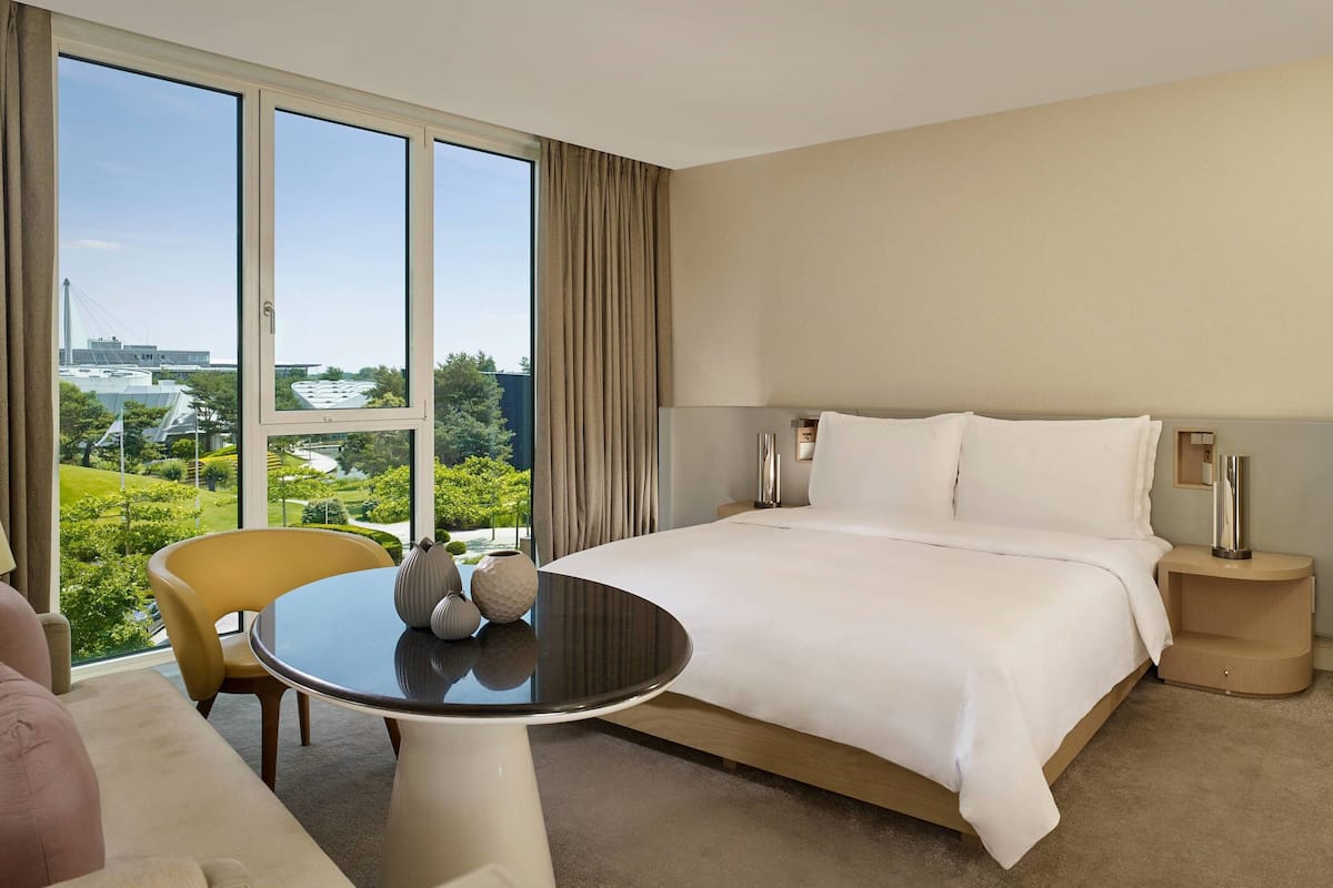 Ein Hotelzimmer mit einem großen Bett, einem runden Tisch mit Dekoration, einem Stuhl und Blick auf Grünflächen und Gebäude.