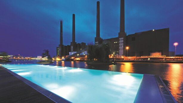 A nighttime view of a swimming pool with a water fountain, surrounded by industrial buildings with tall chimneys. - The Ritz-Carlton, Wolfsburg (Wolfsburg)
