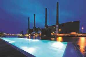 Vista nocturna de una piscina con fuente de agua, rodeada de edificios industriales con chimeneas altas.