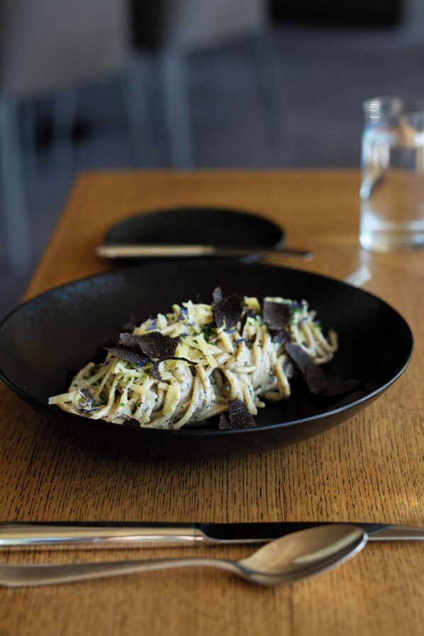 A black bowl of pasta on a wooden table with a spoon and a glass of water in the background.