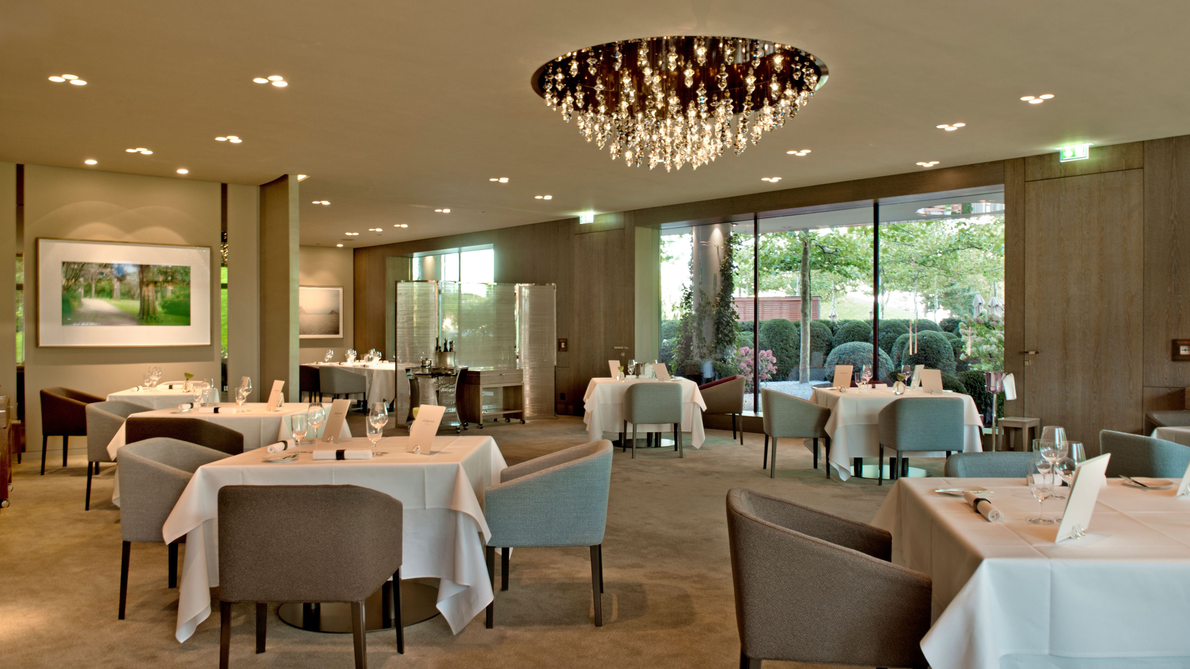 Um restaurante com várias mesas e cadeiras, um candelabro e amplas janelas que oferecem vista para o exterior.