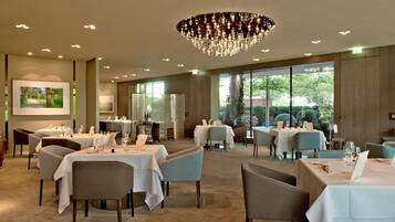 Um restaurante com várias mesas e cadeiras, um candelabro e amplas janelas que oferecem vista para o exterior.