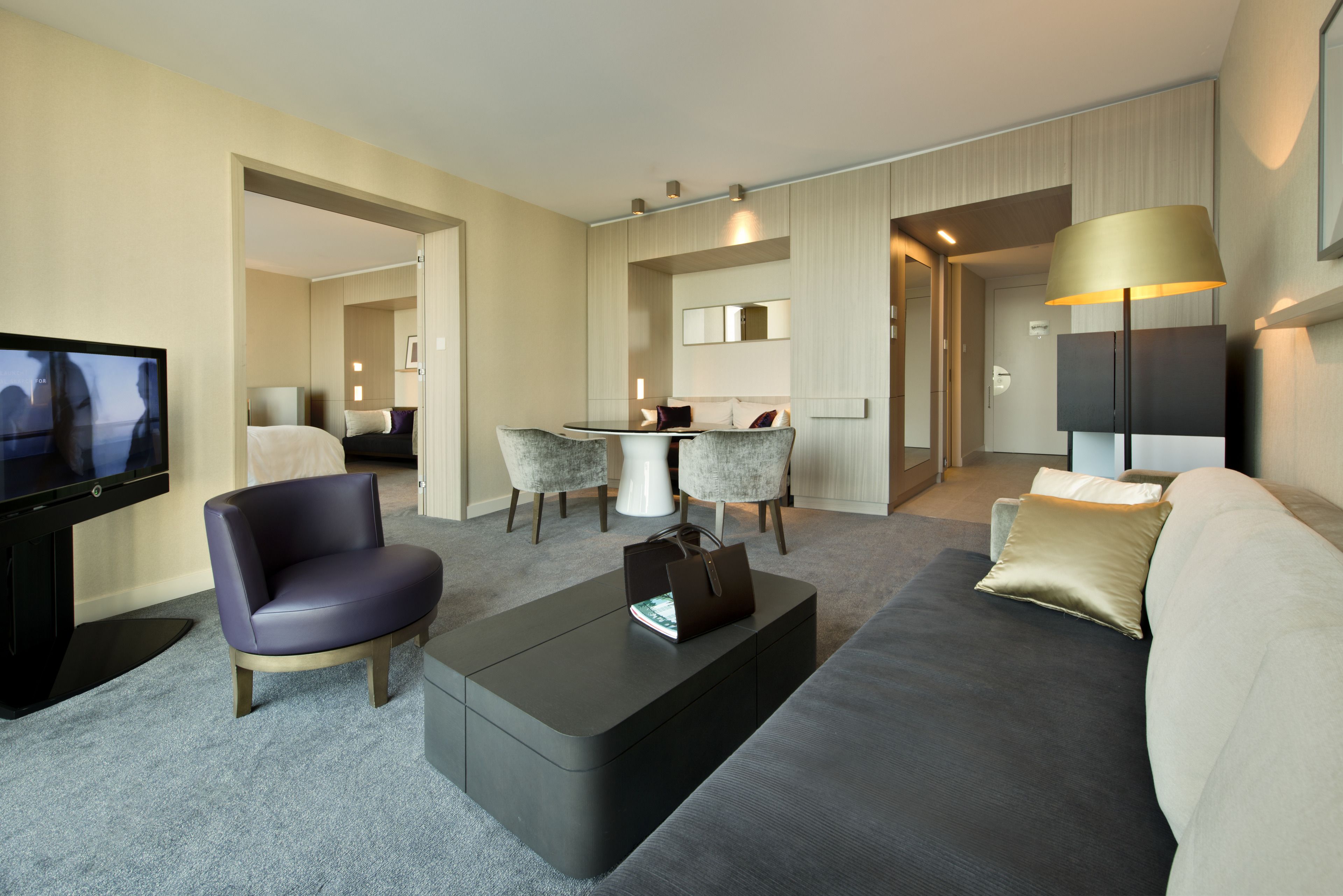 A modern hotel room with a flat-screen TV, a sofa, a coffee table, and a dining area with chairs and a table.