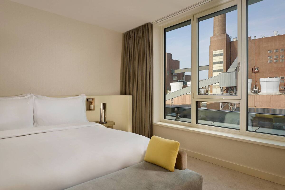 Ein Hotelzimmer mit einem großen Bett, einem Nachttisch, einer Lampe, einem Fenster mit Blick auf ein Gebäude und einem Vorhang.